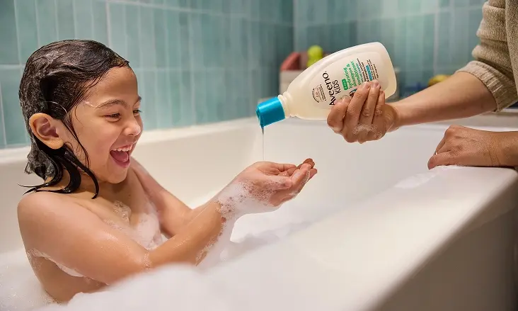 Image of a girl bathing in the tub, her mother helping her with a bottle of Aveeno® Kids Body Bath