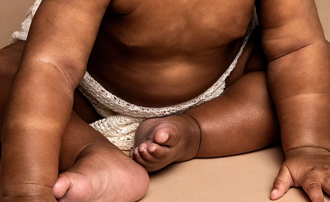 Close up of baby sitting on the floor with legs crossed, showing chest, belly, arms, knees, shins, feet
