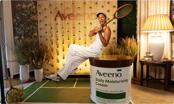 Man grips a tennis racket, positioned in front of a large Aveeno Daily Moisturizing Cream container