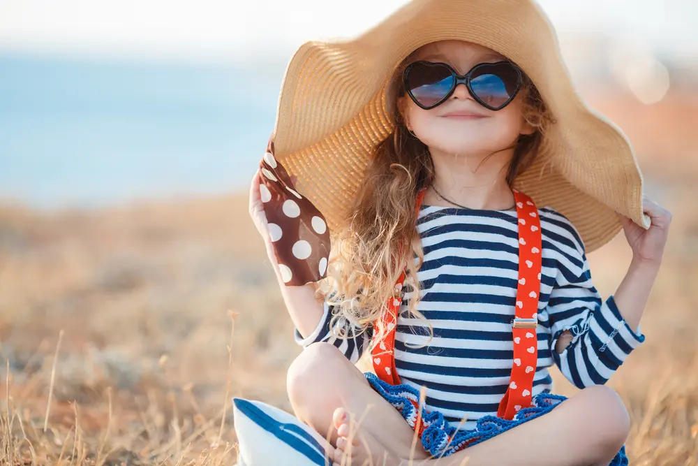 A young girl sits near the beach wearing a sun hat.