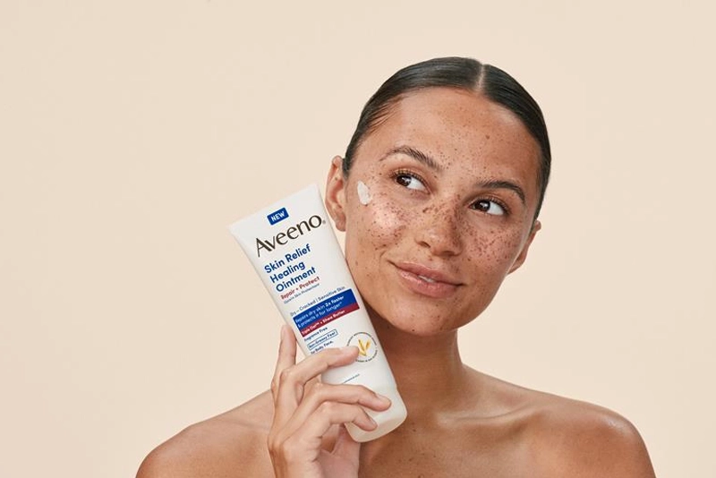 Woman with freckles holds a tube of Aveeno Skin Relief Ointment, showcasing the product application