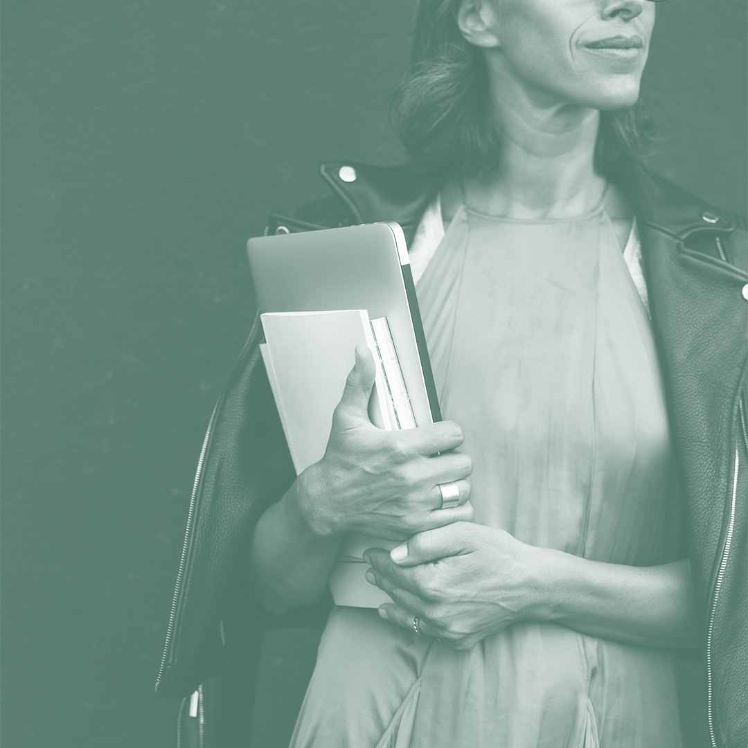 An image of a woman with a leather jacket draped over her shoulder, holding a computer and some notebooks.