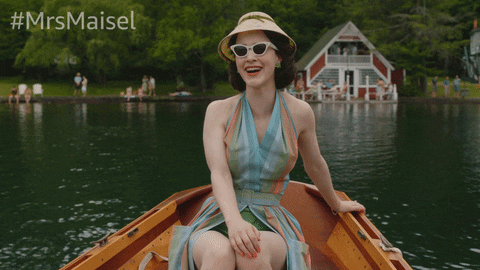 A GIF from ‘The Marvelous Mrs. Maisel’ in which Midge sits in a boat, wearing a bathing suit cover and sunglasses, and laughs.