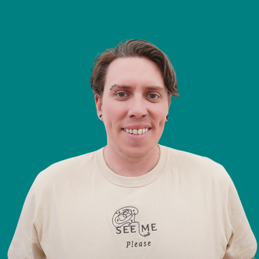 Daniel Leighton, Engagement Lead, he's got wavy short brown hair and eyebrow and nose piercings, smiling in a cream See Me Please embroidered t-shirt