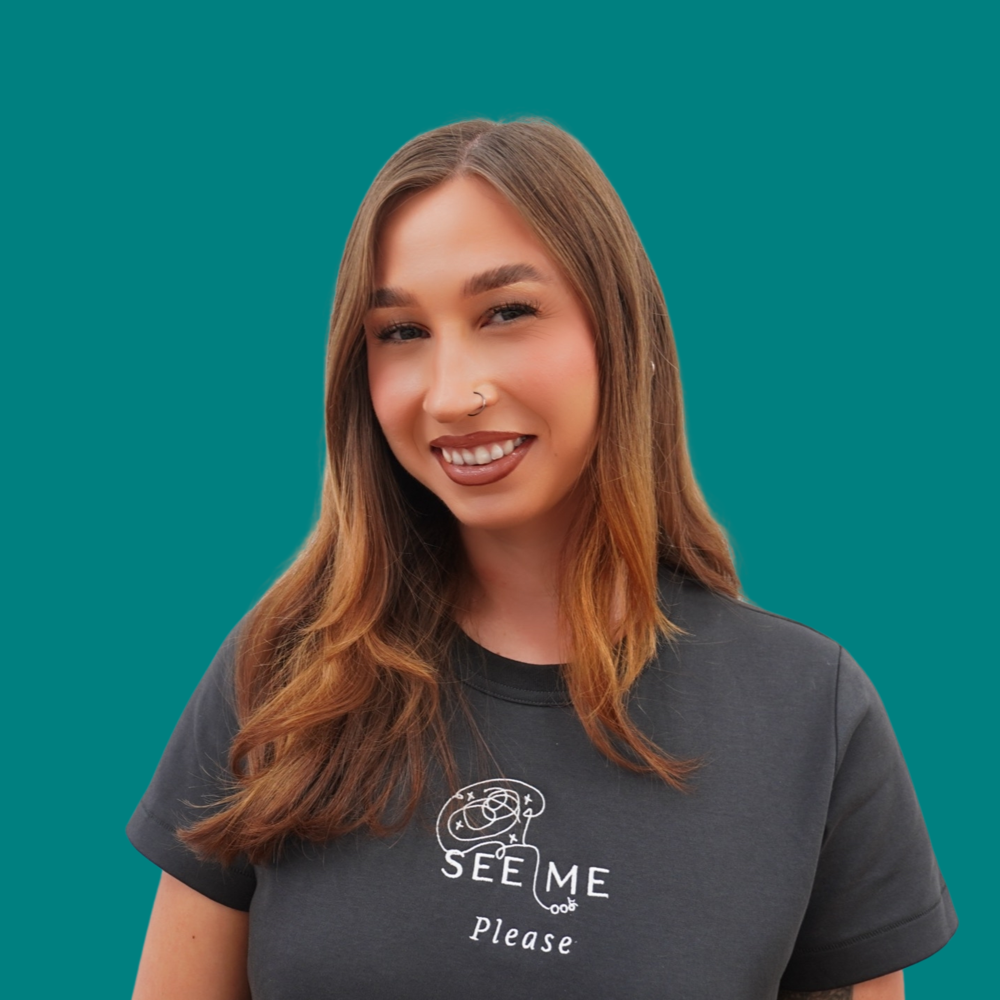 Natalie Laydon, Marketing Specialist, she has long brown hair and a nose ring, smiling in a dark grey See Me Please embroidered t-shirt