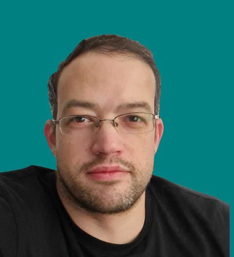 A man with short dark hair, wearing glasses and a black T-shirt, looks directly at the camera with a neutral expression. The background is a solid teal colour.