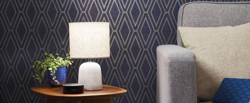 Table lamp switched on and sitting on a wooden table next to a blue plant pot and Amazon speaker with a grey sofa next to the table and a diamond-patterned blue and gold wallpaper in the background