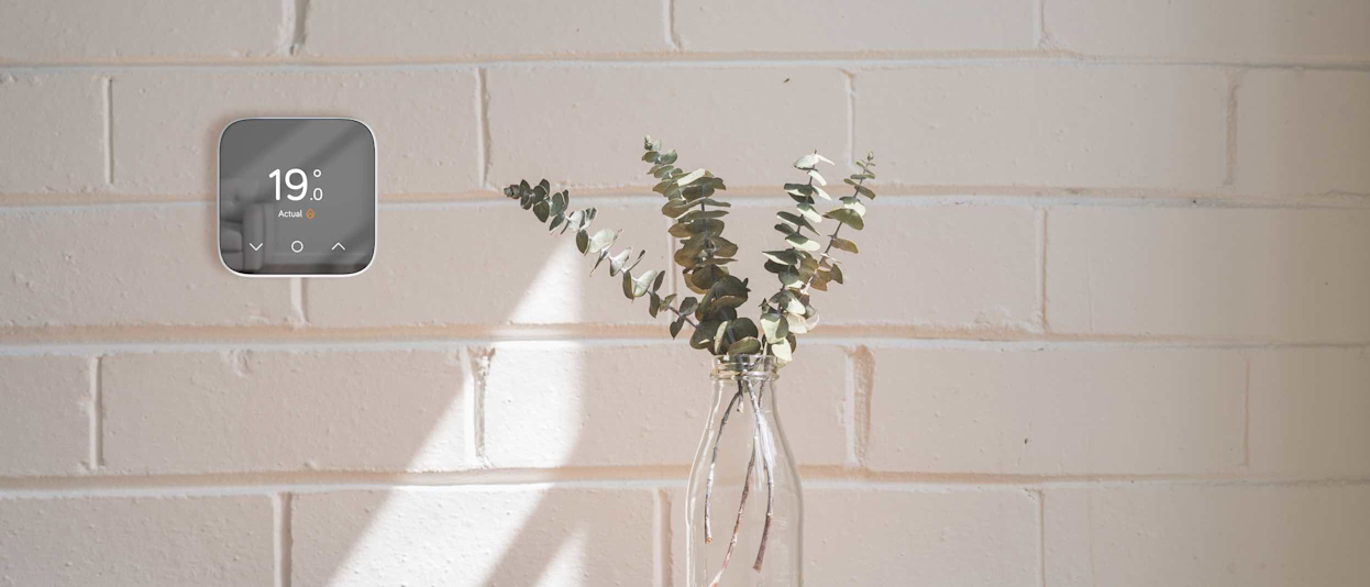 Hive Thermostat Mini attached to a white interior brick wall, with a clear bottle holding green leaves sitting on a wooden table underneath