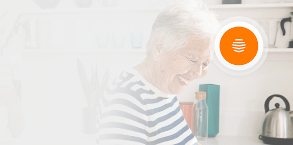 An older woman laughing and holding a saucepan over a gas hob, with a silver kettle sitting on a kitchen counter in the background, and a Hive logo superimposed in the top right of the image