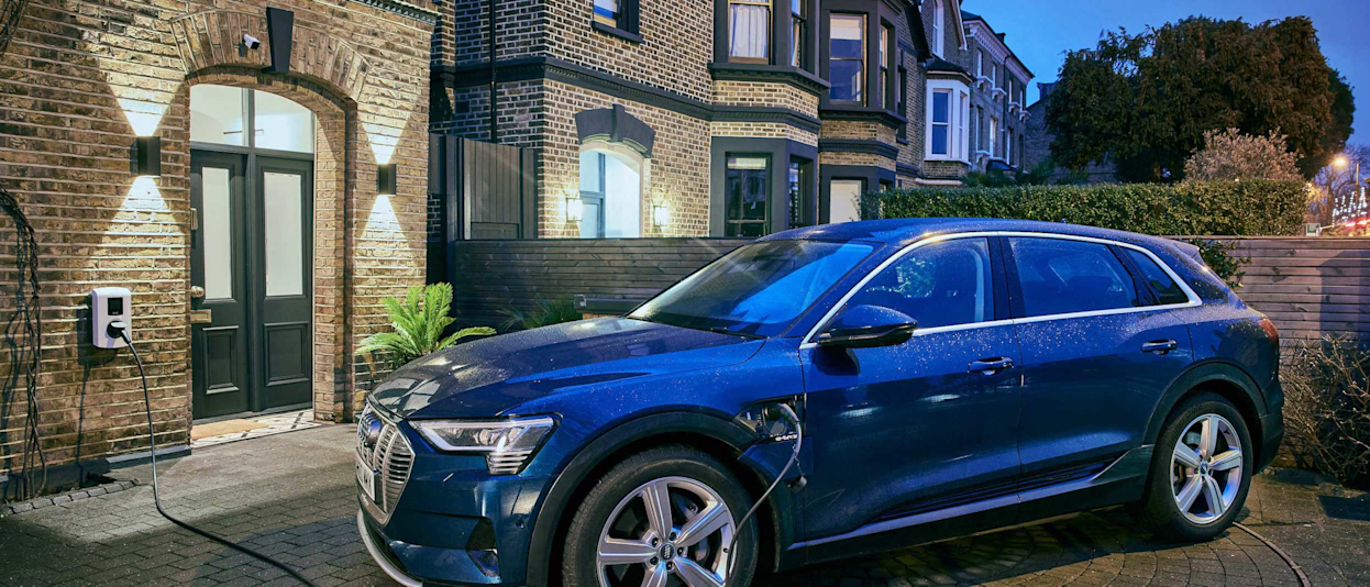 Dark blue car sitting in front of a brown brick house with an EV Charger on the wall of the house and a cable attached from the charger to the car