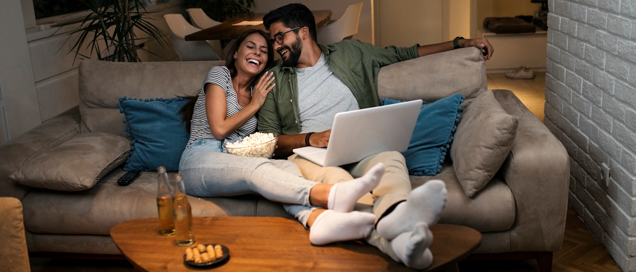Laughing young couple sitting on a brown suede sofa with a laptop and a bowl of popcorn on their laps, and their feet resting on a wooden coffee table