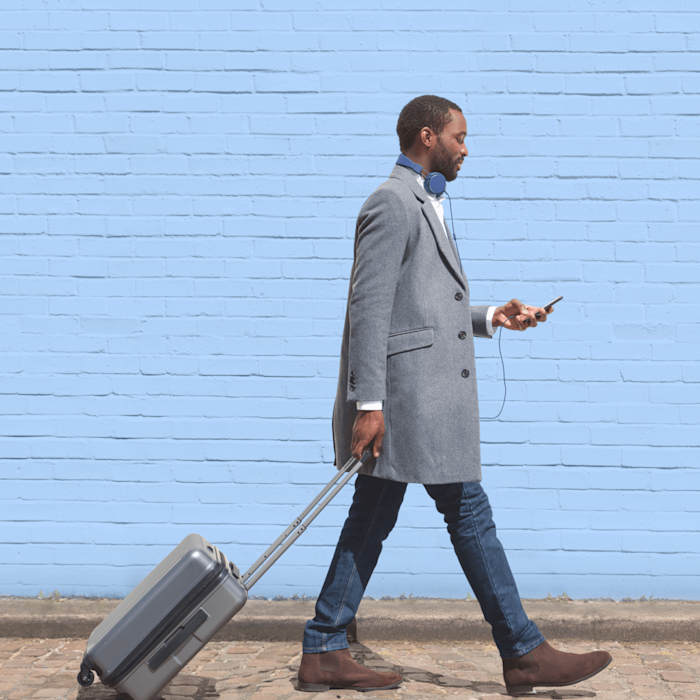 Man in smart casual clothing walking in front of a blue brick wall looking at his phone and pulling a suitcase behind him