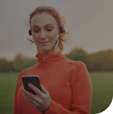 Woman in an orange running top and wearing black earphones looking at a smartphone in her hand with a row of trees in the background