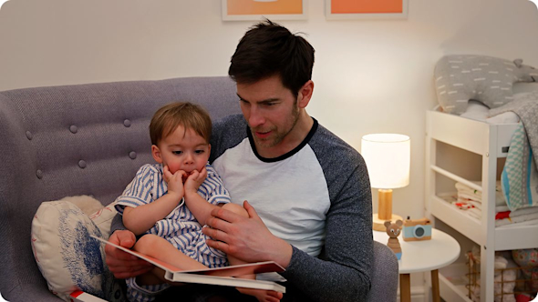 Young dad sitting on a grey armchair with his son in his lap reading a book with a lamp behind them on a white table and a small bed to the right of the room