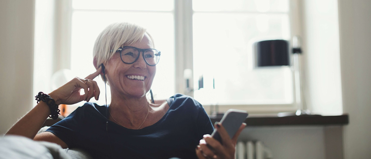 Mature woman sitting on a sofa at home talking on a phone with earphones on