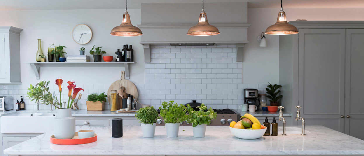 Bright white kitchen with three copper lights hanging from the ceiling, and plant pots with green herbs, a bowl of fruit and a vase filled with red flowers sitting on the kitchen island