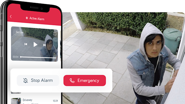 Side angle of smartphone showing Hive app with a pop-out icon showing a red emergency graphic, with a young man in a hoodie trying to get in the front door of a house and looking into a security camera