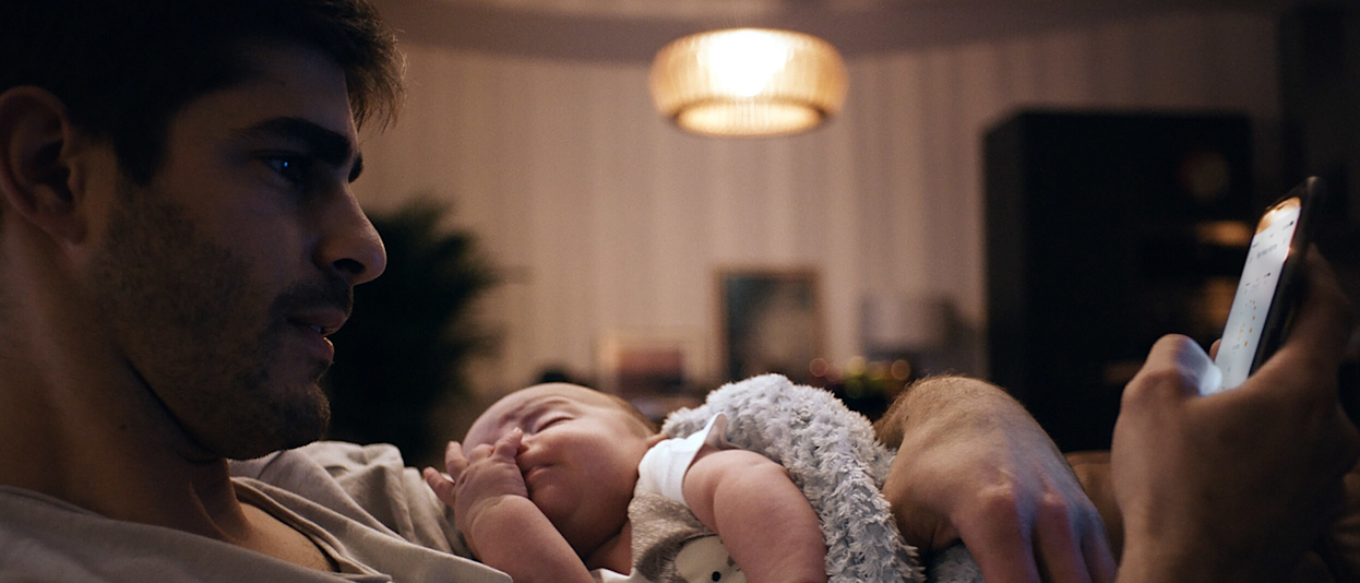 Young dad looking at his smartphone while holding a small baby in his arms with a bright ceiling light and stripy wallpaper in the background