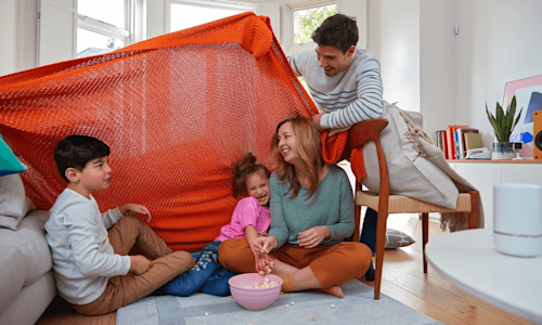 Family playing in a bright living room with an orange blanket being used to cover a fort made from the sofa and a dining room chair