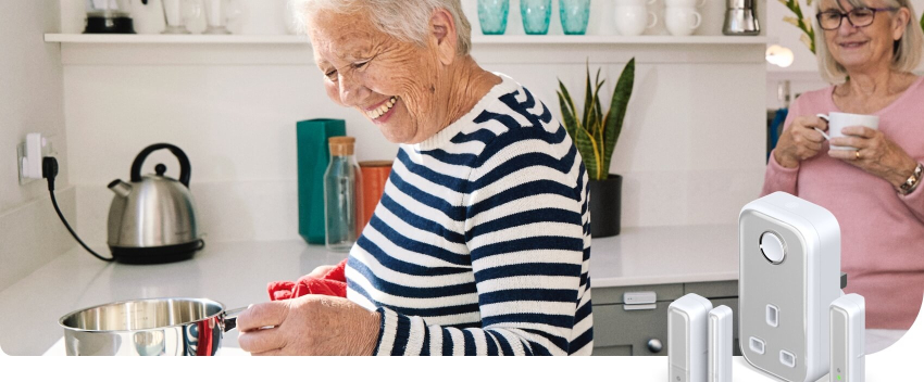 Two older woman laughing in a bright white kitchen with one woman holding a mug and the other woman holding a saucepan over a gas hob, a shelf in the background shows blue glasses and white mugs, and in the bottom right corner there are Hive smart devices superimposed over the image