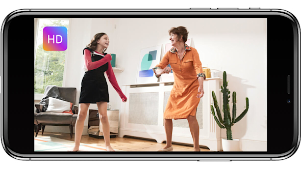 Smartphone screen showing a girl and an older woman dancing in a living room with abstract art on the walls behind them and a Hive Motion Sensor attached in the corner of the room, with HD logo superimposed in the top left corner