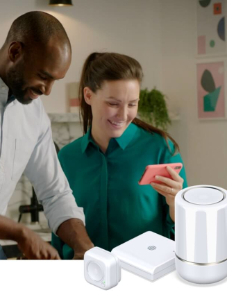 Couple in a kitchen looking at a pink smartphone and smiling with plants in the background against a white marble wall, and Hive smart devices superimposed in the bottom right corner