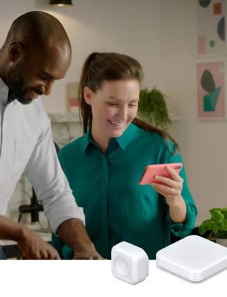 Couple in a kitchen looking at a pink smartphone and smiling with plants in the background against a white marble wall, and Hive smart devices superimposed in the bottom right corner