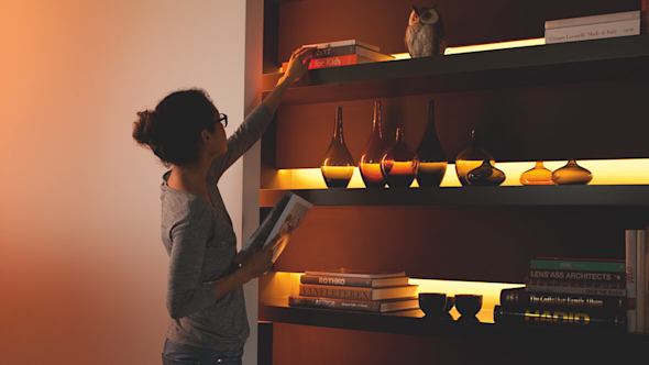 Woman reaching up to a pile of books on a large wooden bookcase with bright strips of light at the back of each shelf, with books and vases on the lower shelves
