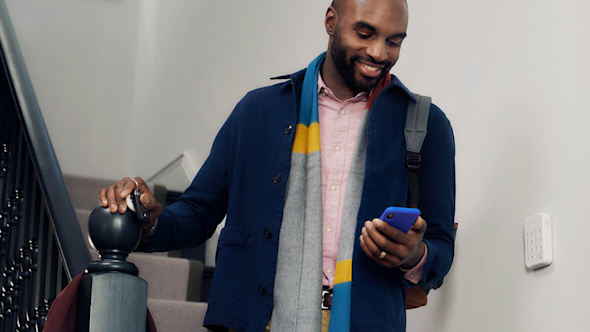 Smiling man wearing a navy jacket and brightly coloured scarf walking down the stairs while looking at a smartphone with a Hive Keypad attached to the wall on the right