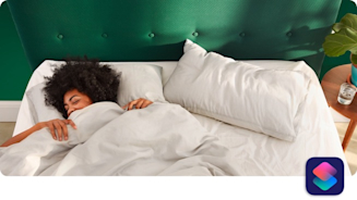 Woman, lying in a bed with a green headboard, pulling a white duvet up to her face with a wooden bedside table to the right of the bed with a glass of water and a plant on it