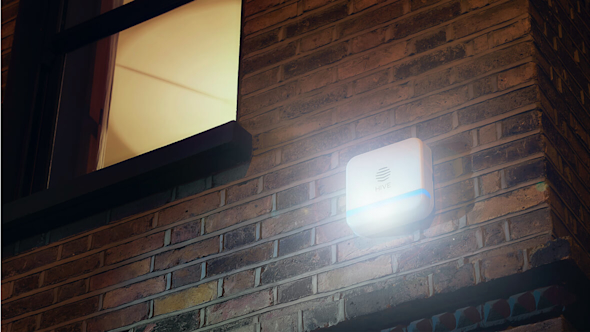 Hive Siren attached to a brown brick exterior wall of a house, at night, with a light showing at the bottom of the siren, and a window with a light on in the top left