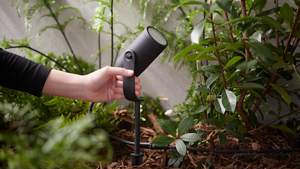 A hand positioning a Philips Hue Lily Outdoor Smart Light that is sat among a range of green plants and bushes