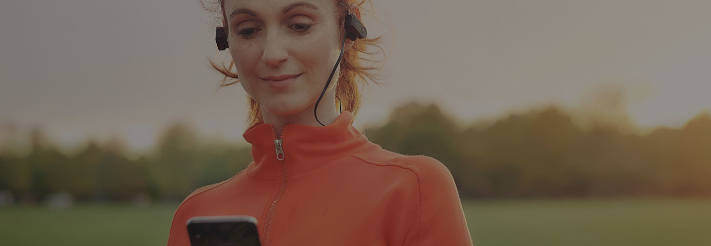 Woman in an orange running top and wearing black earphones looking at a smartphone in her hand with a row of trees in the background