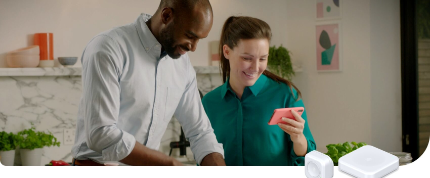 Couple in a kitchen looking at a pink smartphone and smiling with plants in the background against a white marble wall, and Hive smart devices superimposed in the bottom right corner