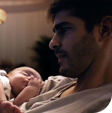 Young dad looking at his smartphone while holding a small baby in his arms with a bright ceiling light and stripy wallpaper in the background