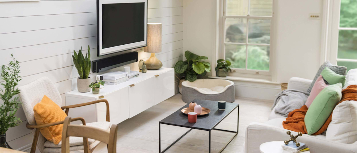 White and cream living room with TV on the wall, and a coffee table, sofa and armchair around the room with sash windows looking out to a garden in the background