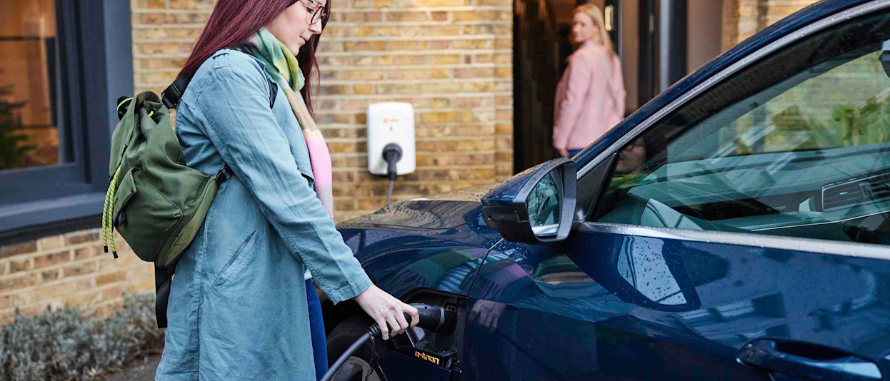 Woman in a blue jacket and wearing a green backpack is charging her car with electric power in front of a brown brick house using a Hive EV Charger attached to the wall
