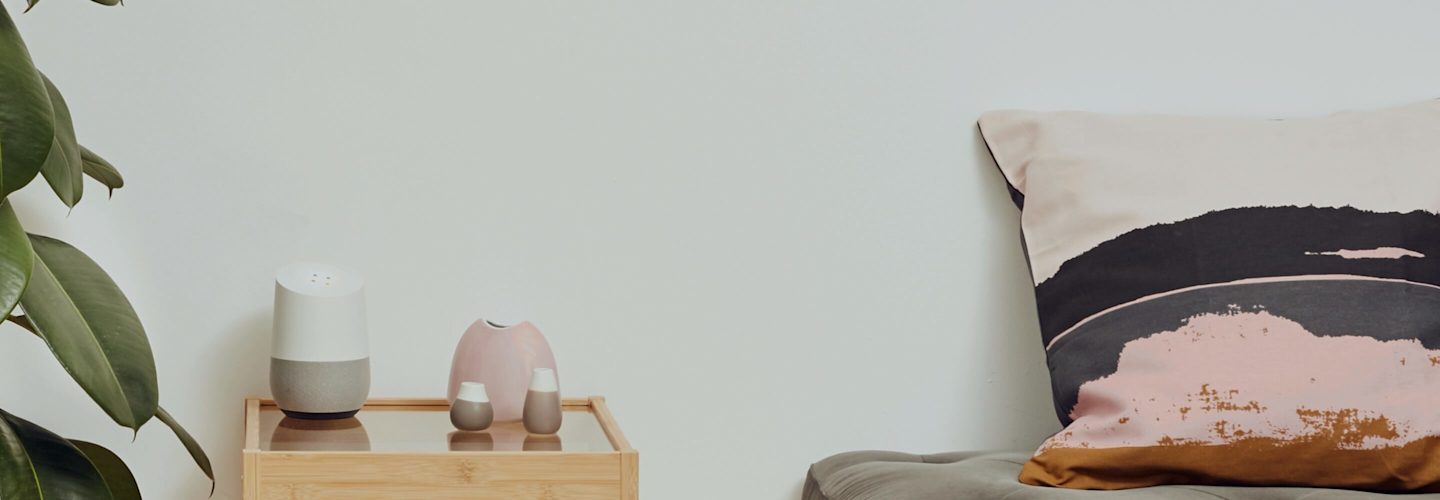 Google Speaker and ceramic pots sitting on a glass and wooden unit next to a tall green plant on the left with a cream, dark grey and pink cushion to the right and a grey painted wall in the background