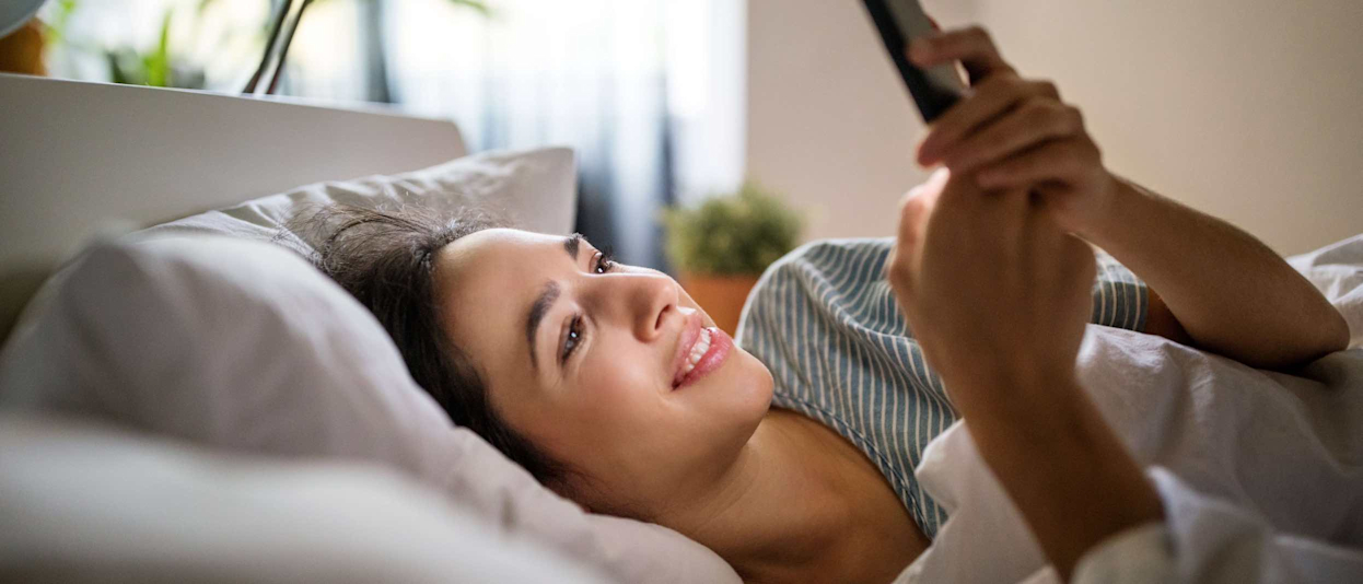 Woman in bed looking at her smartphone with a window in the background and plants on the windowsill