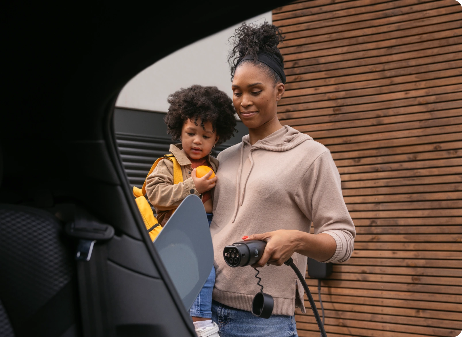 A person holding an EV charging cable while carrying a child beside a parked electric vehicle outside a home.