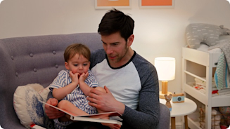 Young dad sitting on a grey armchair with his son in his lap reading a book with a lamp behind them on a white table and a small bed to the right of the room