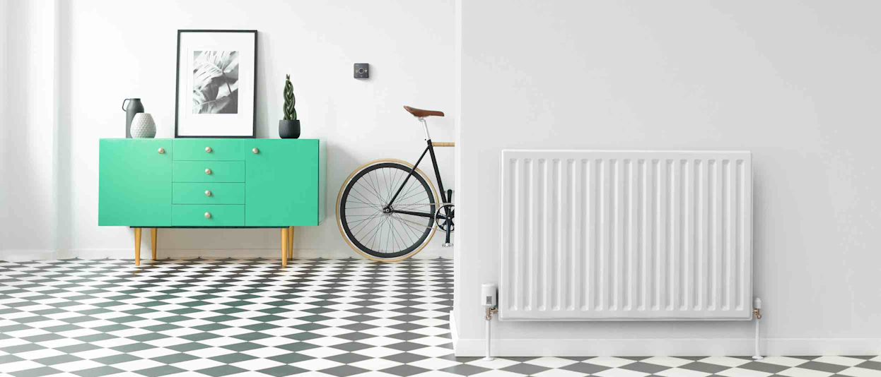 Large hallway with black and white floor tiles, a radiator in the foreground wall with a Hive Radiator Valve attached, a green drawer unit in the background next to a Hive Thermostat on the wall and a bike propped up against the wall
