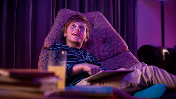 Boy sitting on a modern grey armchair while using a tablet, with curtains in the background, and books and a glass of juice in the foreground, with a purple light lighting up the space