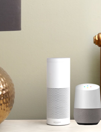 Large gold lamp with dark grey lampshade on a white unit next to an Amazon smart speaker and a Google smart speaker, with an old-style copper radiator in the background