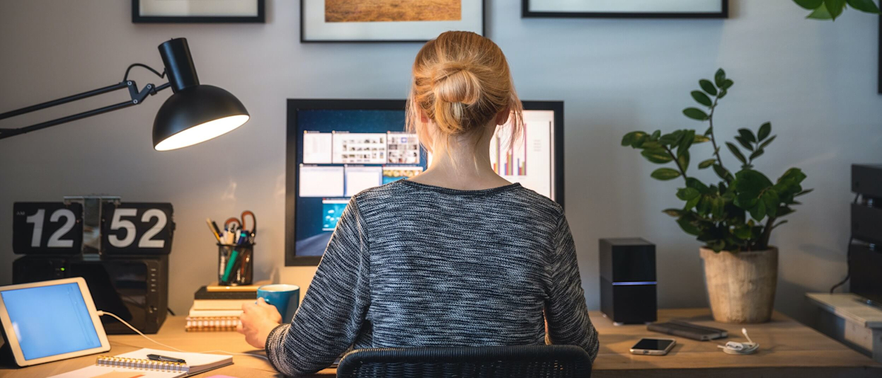 Woman sitting, with her back to the camera, working on a computer at a wooden desk with a big number clock, speakers, tablet, plants and pencil holder on the desk