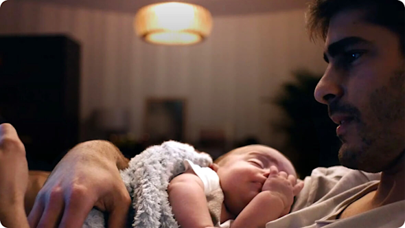A man cradling his child in a dimly lit room while looking at his smartphone