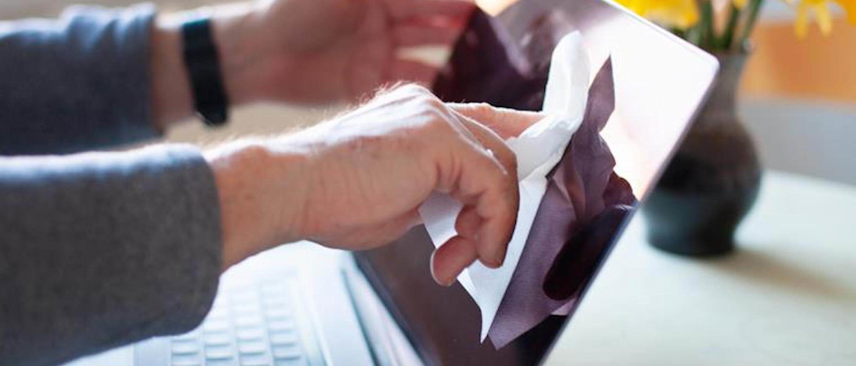 Close-up of a man's hands cleaning a laptop screen with a white tissue with a green vase of yellow flowers in the background