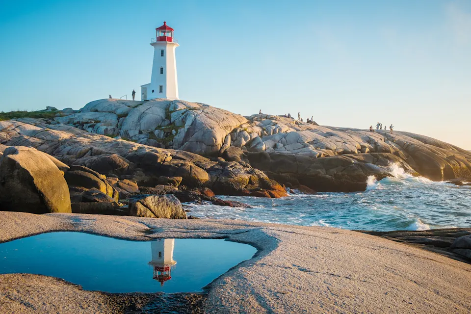 Peggy's Cove Lighthouse in Nova Scotia reflected in a tidal pool on a rocky shoreline during sunset.
