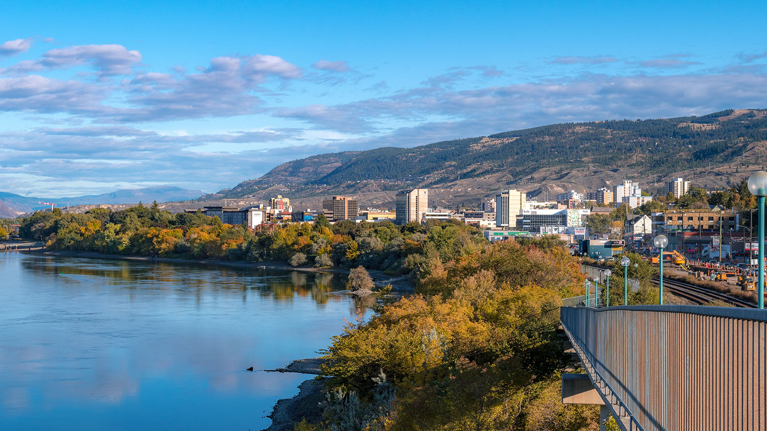 View of Kamloops cityscape with a river, greenery, and mountains in the background on a partly cloudy day.