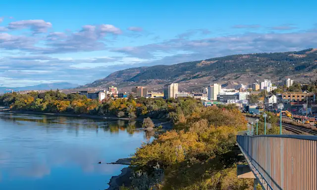 View of Kamloops cityscape with a river, greenery, and mountains in the background on a partly cloudy day.